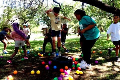 Children playing an outdoor game with colorful balls and a tire in a grassy area with trees.