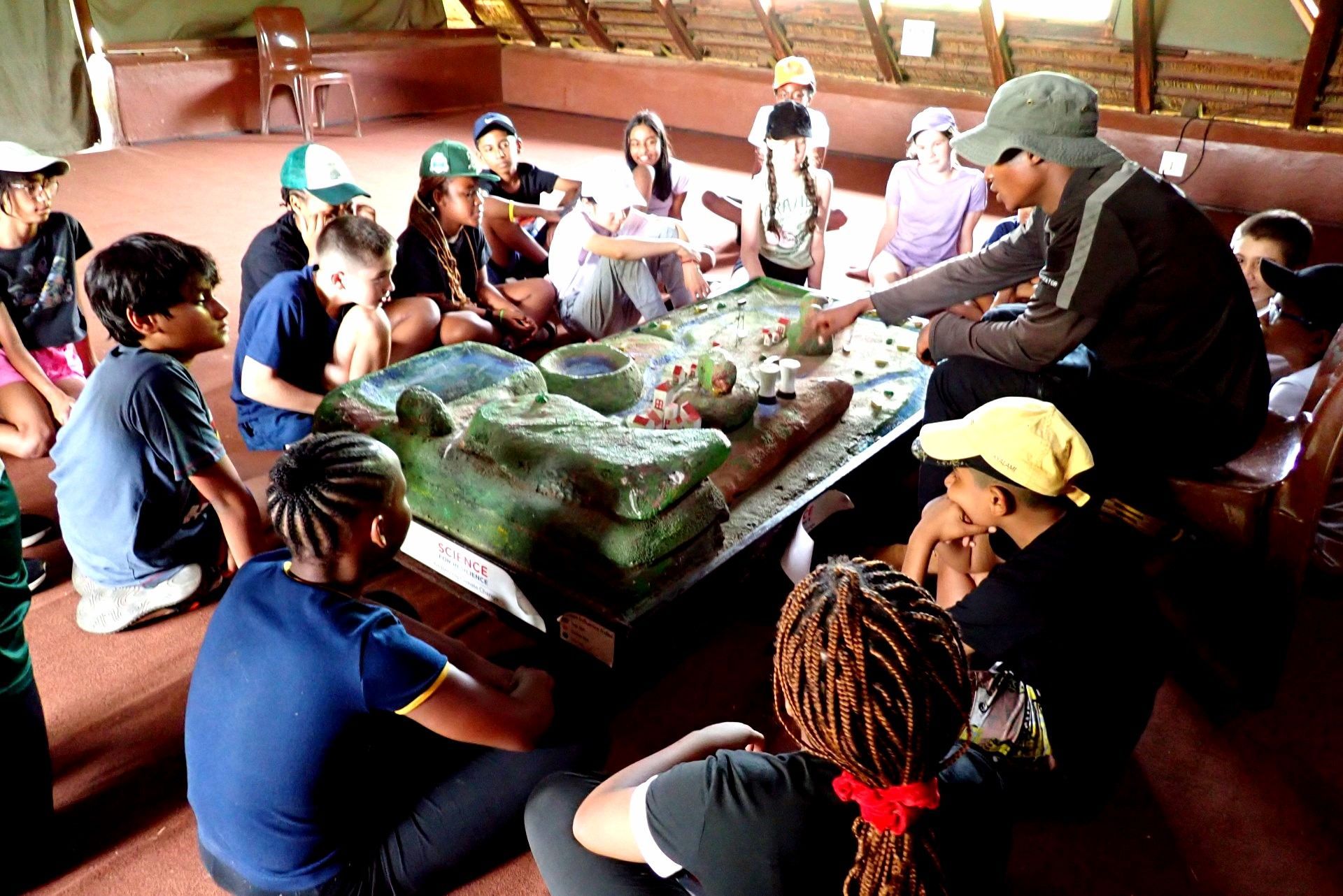 Group of children gathered around a table with a topographical model, a guide pointing to it inside a building.