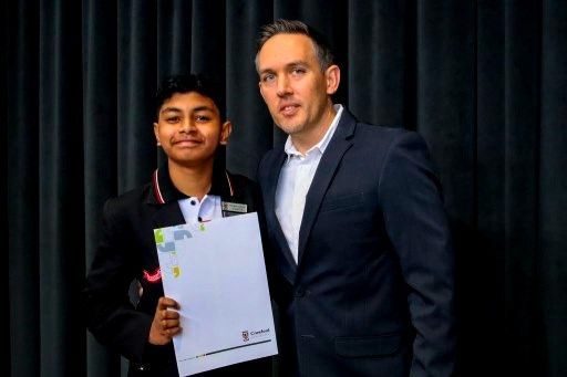Boy in school uniform and man in a suit, both smiling, holding a certificate in front of a dark backdrop.