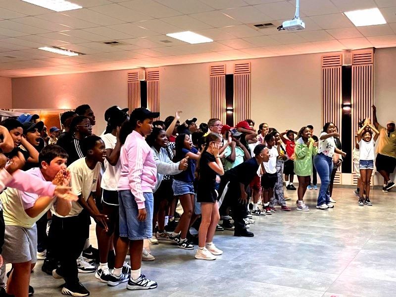 Group of people in a large room, many with hands raised, looking upward, possibly at a screen.