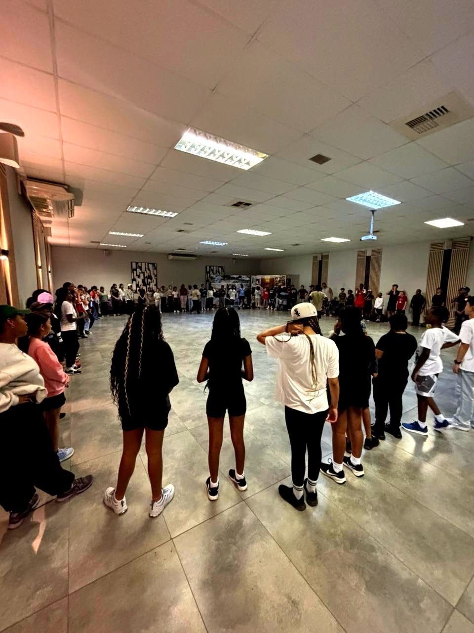 People in a large room, likely a dance or performance practice. Some are in lines, others seated; soft lighting.
