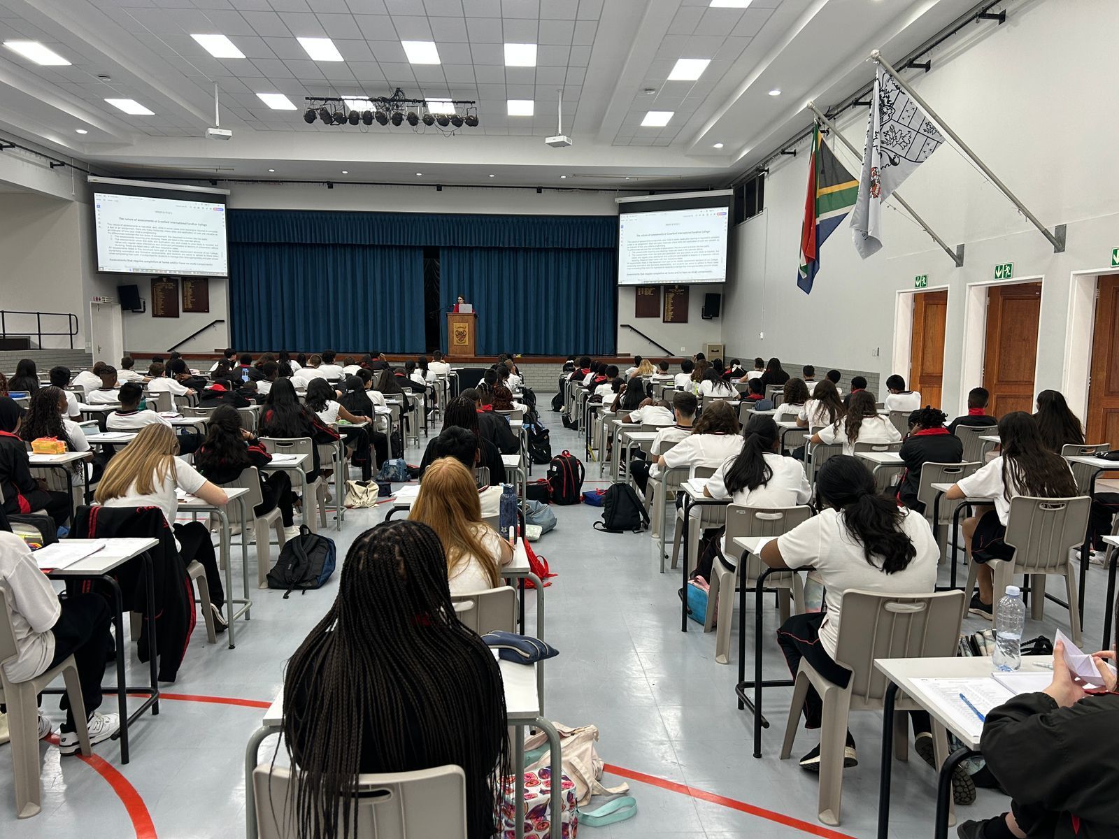 Large room, students seated at desks taking a test. Instructor stands on stage, South African flag displayed.