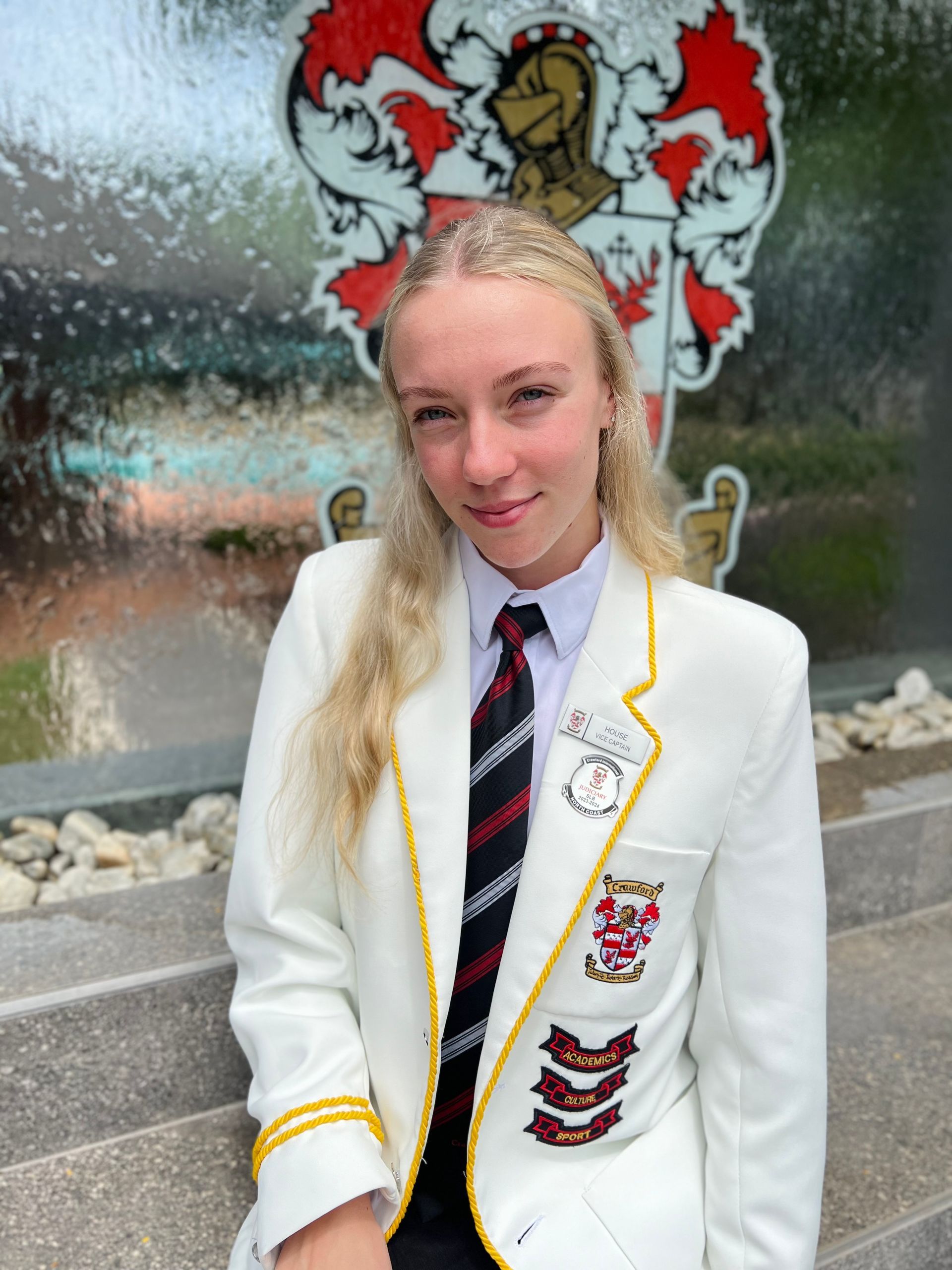 A girl wearing a white jacket and tie is standing in front of a wall with a coat of arms on it.