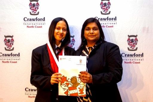 Woman and girl holding certificate, in front of a school banner. Both are smiling, the girl is wearing a red sash.