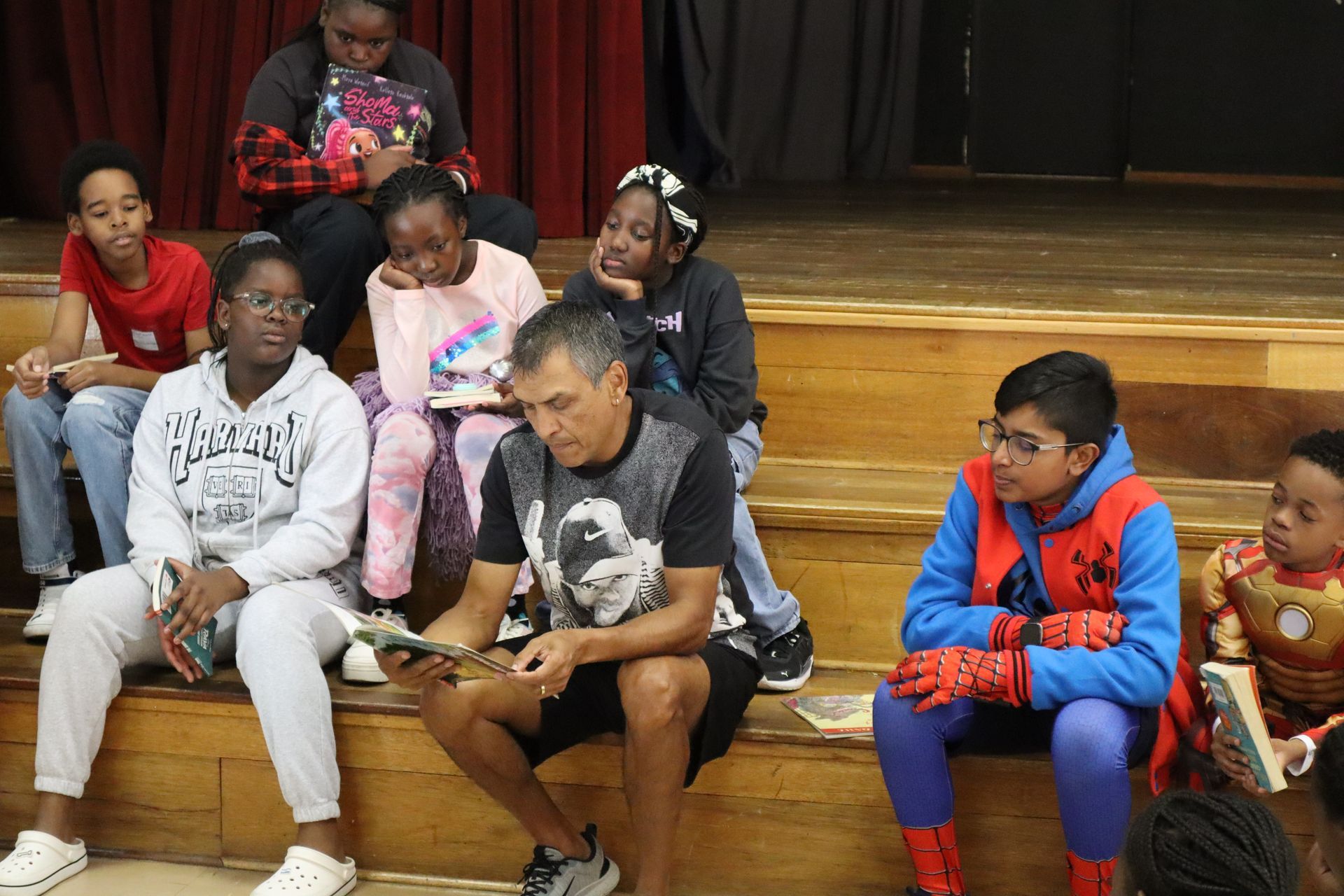 A group of children are sitting on a stage watching a man in a spiderman costume read a book.
