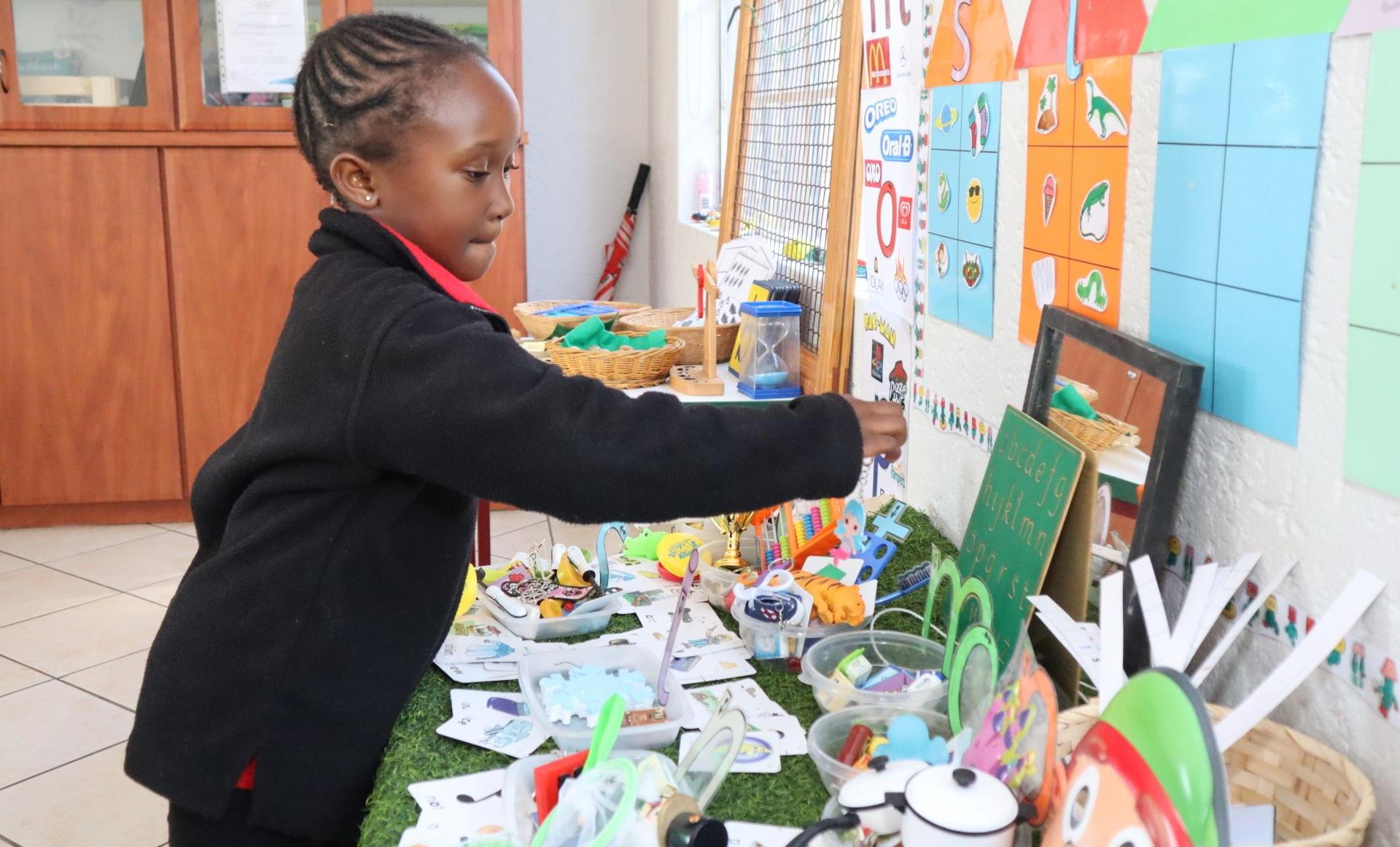 Girl reaching for an item on a table in a classroom with colorful posters and supplies.