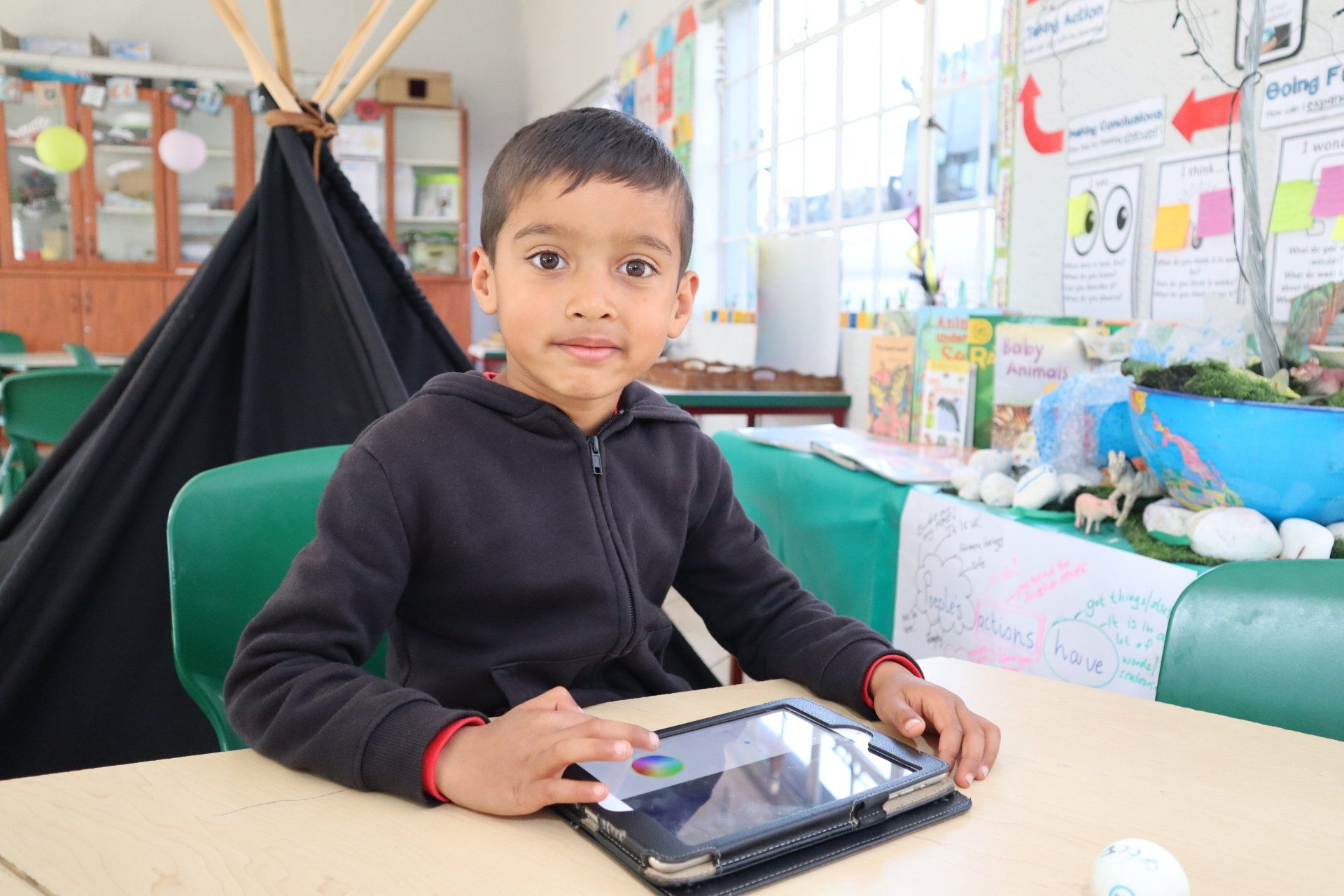Boy using a tablet at a table in a classroom with a teepee, art supplies, and colorful displays.