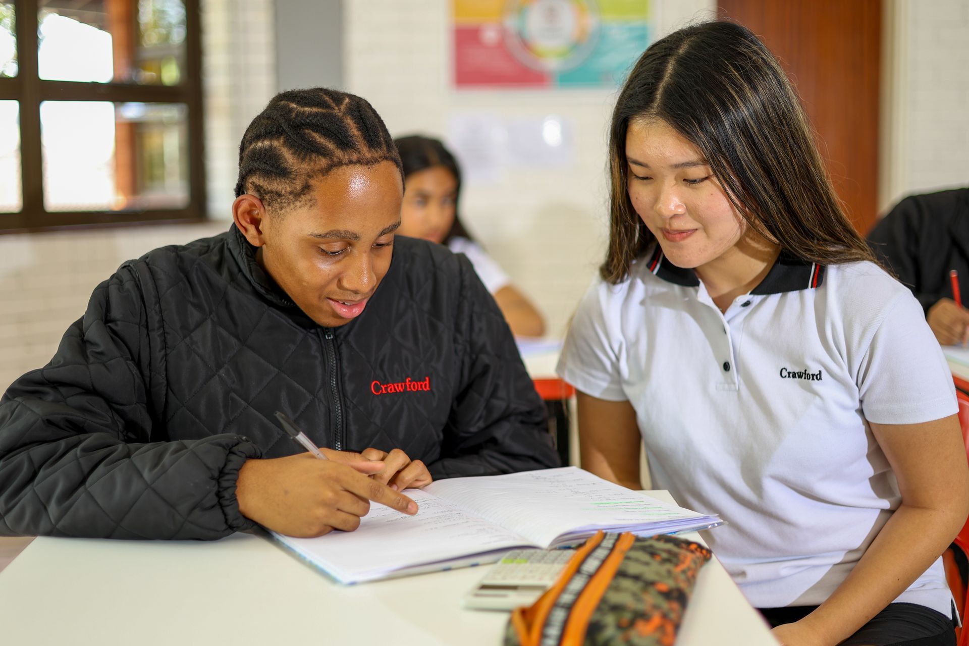 Two students, one with braids, work together at a desk, looking at a notebook.