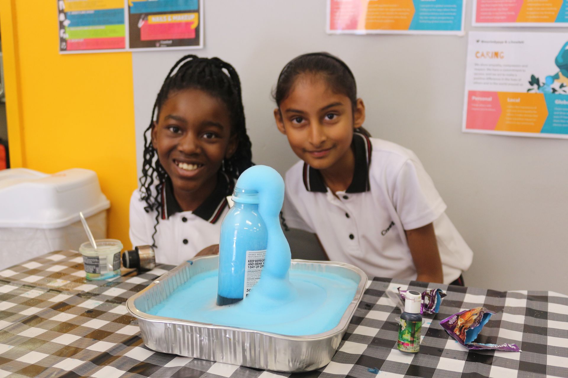 Two young girls are sitting at a table with a container of blue liquid.