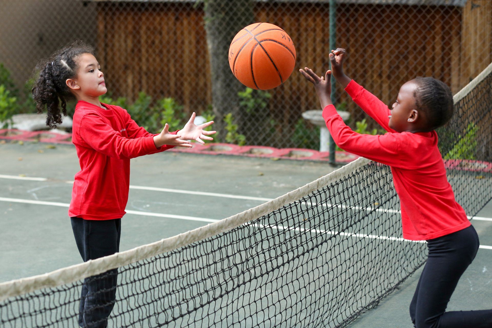 Two children in red shirts play basketball over a net on a green court.
