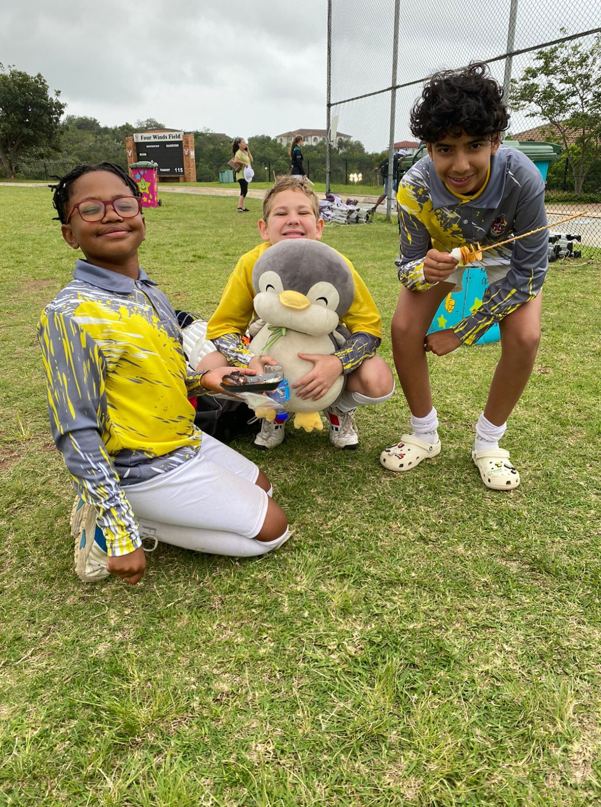 Three children are kneeling in the grass holding a stuffed penguin.