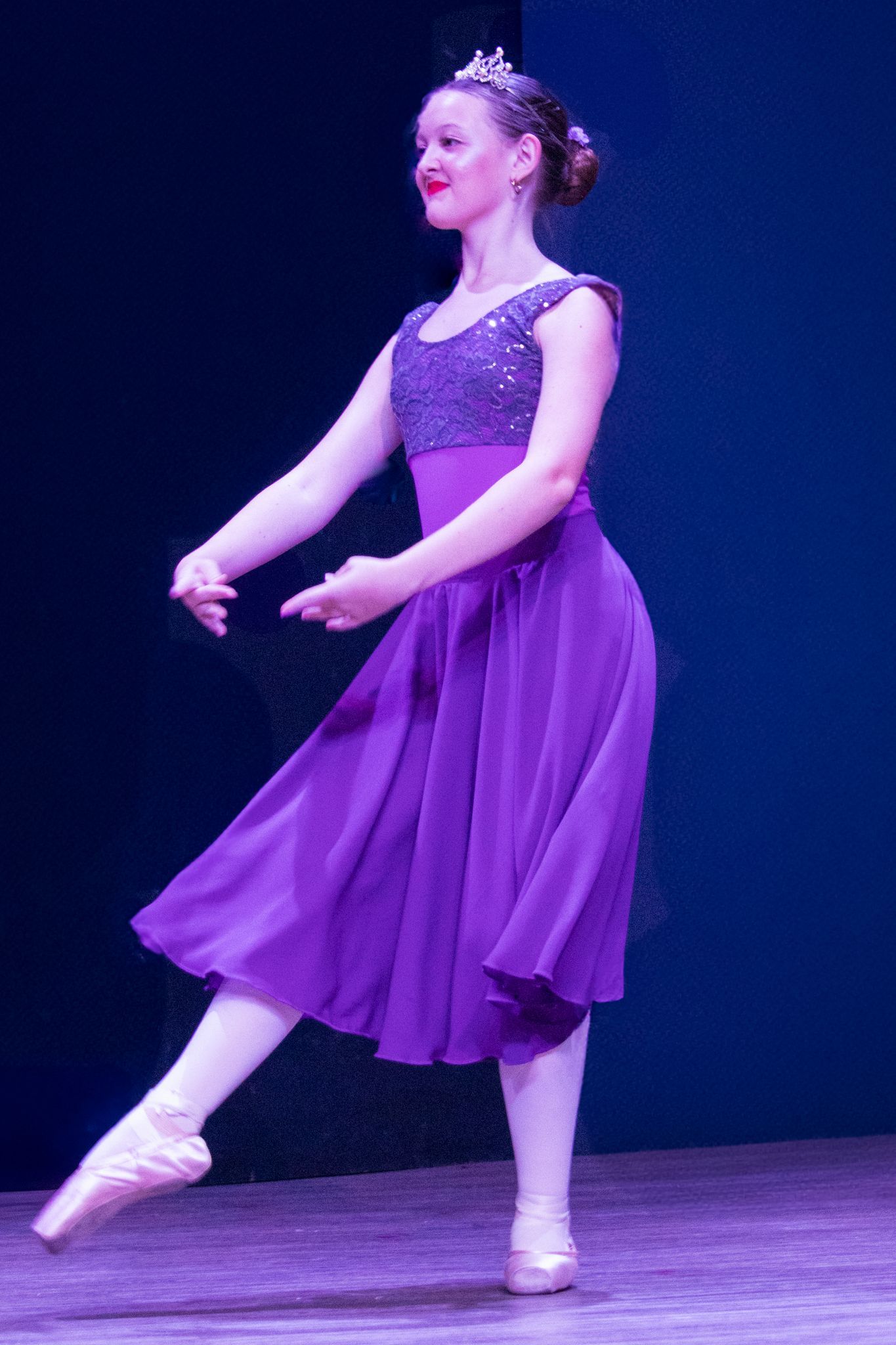 Ballerina in purple dress and tiara, en pointe on stage, arms gracefully extended.
