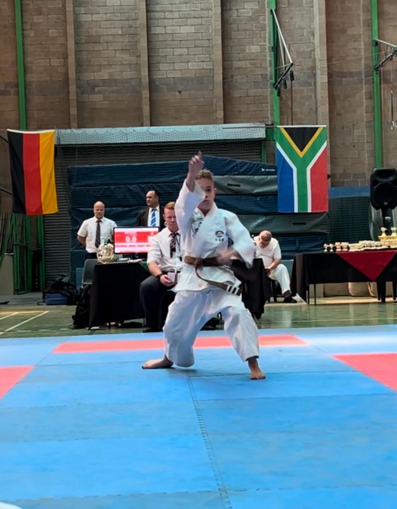 Karate competitor performing a move on a blue mat, flags of Germany and South Africa in background.