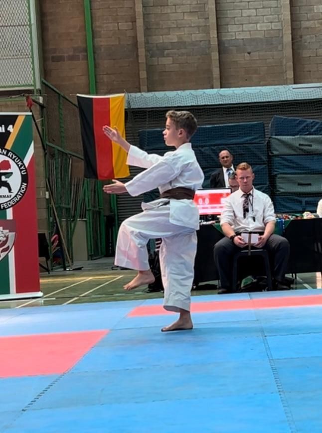 Young person in karate uniform performs a stance on a mat, German flag in the background.
