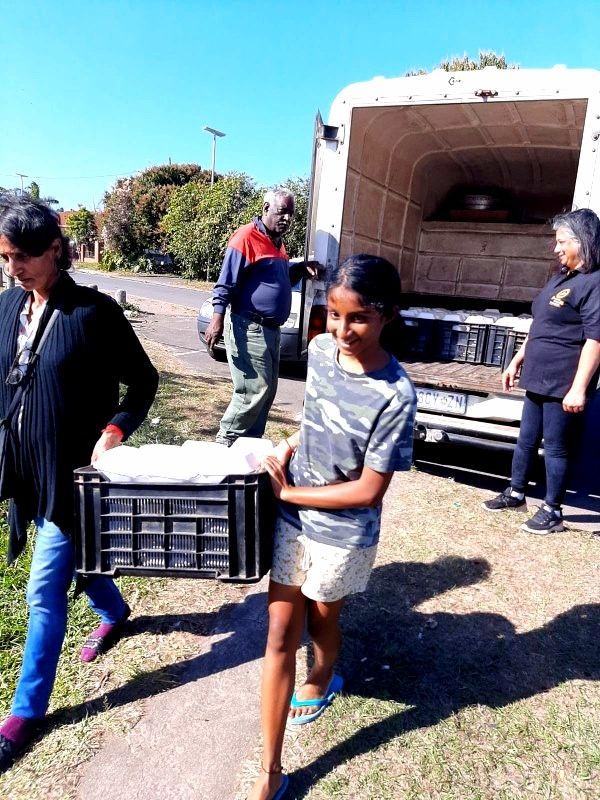 People unloading food from a truck. A person carries a crate, smiling. Outdoors, sunny.