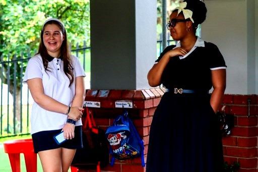 Two women standing outside, one smiling in a white shirt and dark shorts, the other in a black dress, both looking to the side.