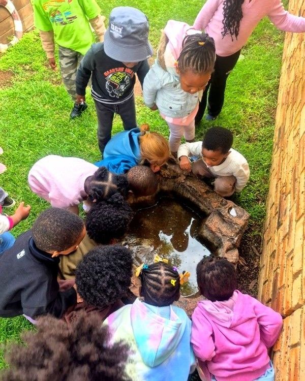 Children gather around a puddle, looking down. Grass and a brick wall are in the background.
