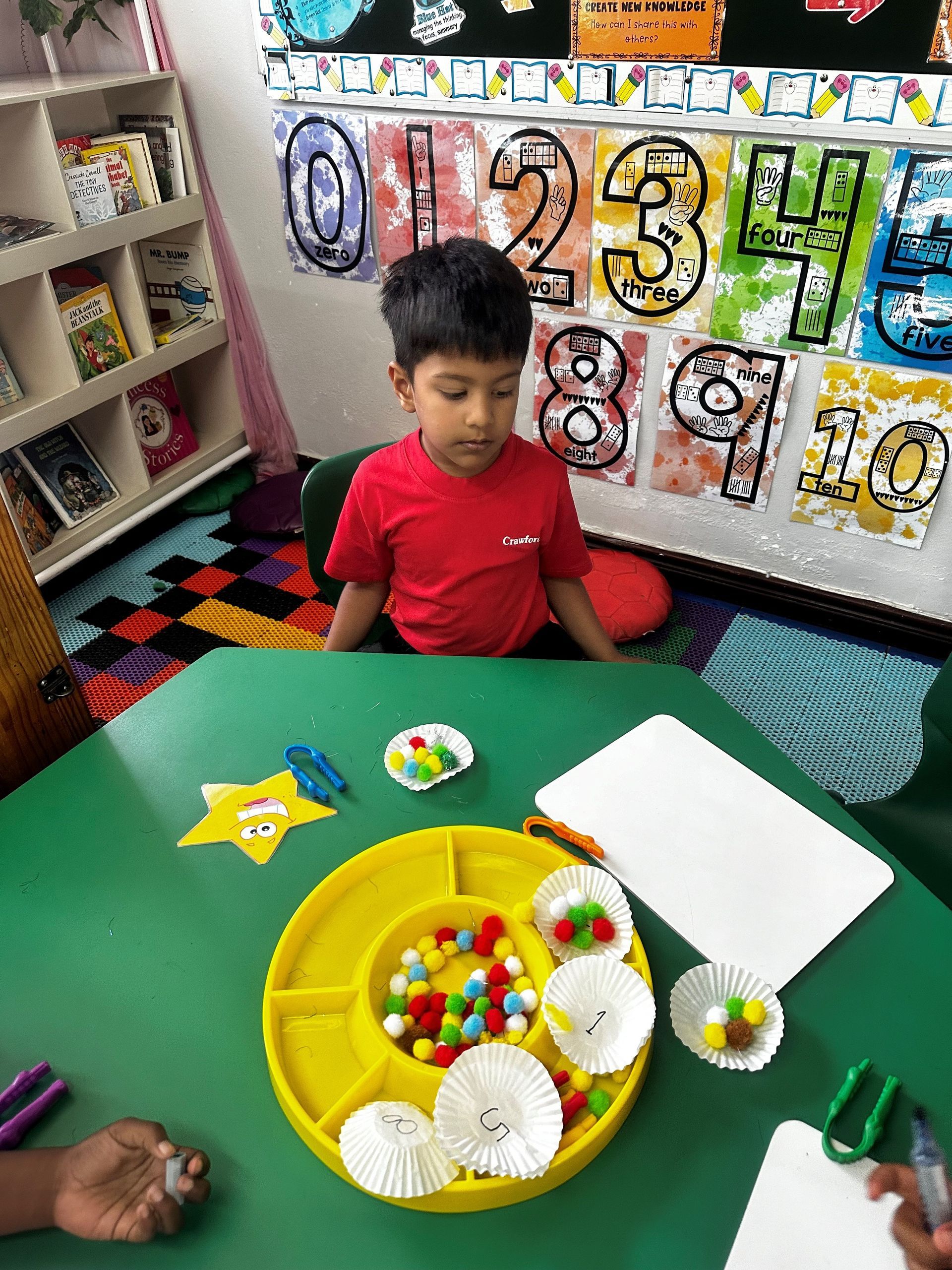A young boy is sitting at a table with a tray of pom poms.
