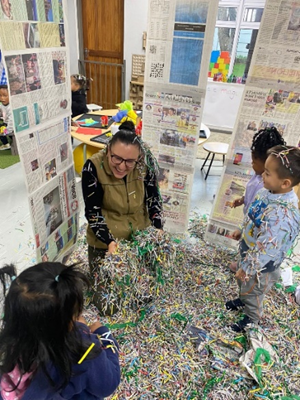 A woman is standing next to a pile of shredded paper in a room with children.