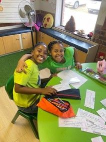 Two young girls are sitting at a green table in a classroom.