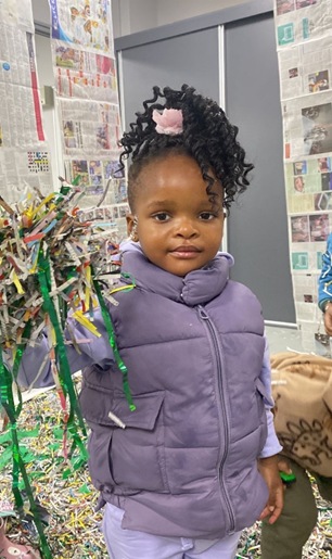 A little girl in a purple vest is standing in front of a pile of shredded paper.