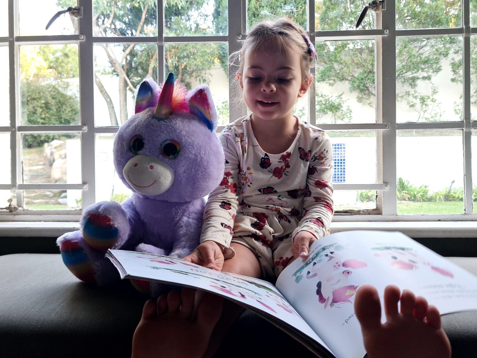 A little girl is reading a book next to a stuffed unicorn