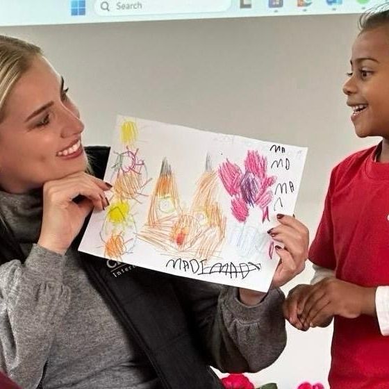 Woman smiling, holding child's drawing with colorful flowers; child smiles too, wearing a red shirt.