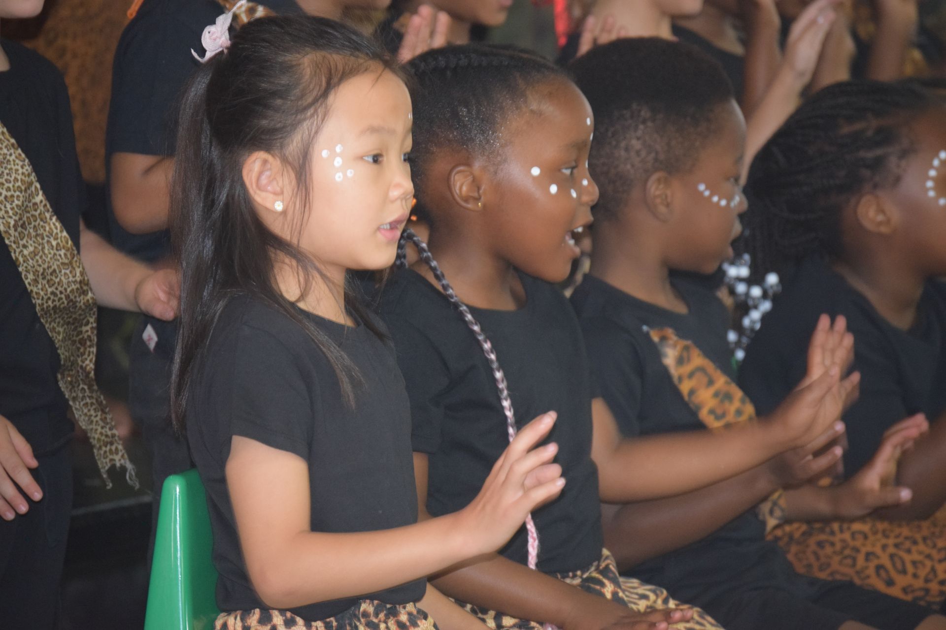 A group of children are sitting in a row wearing black shirts and leopard print skirts.