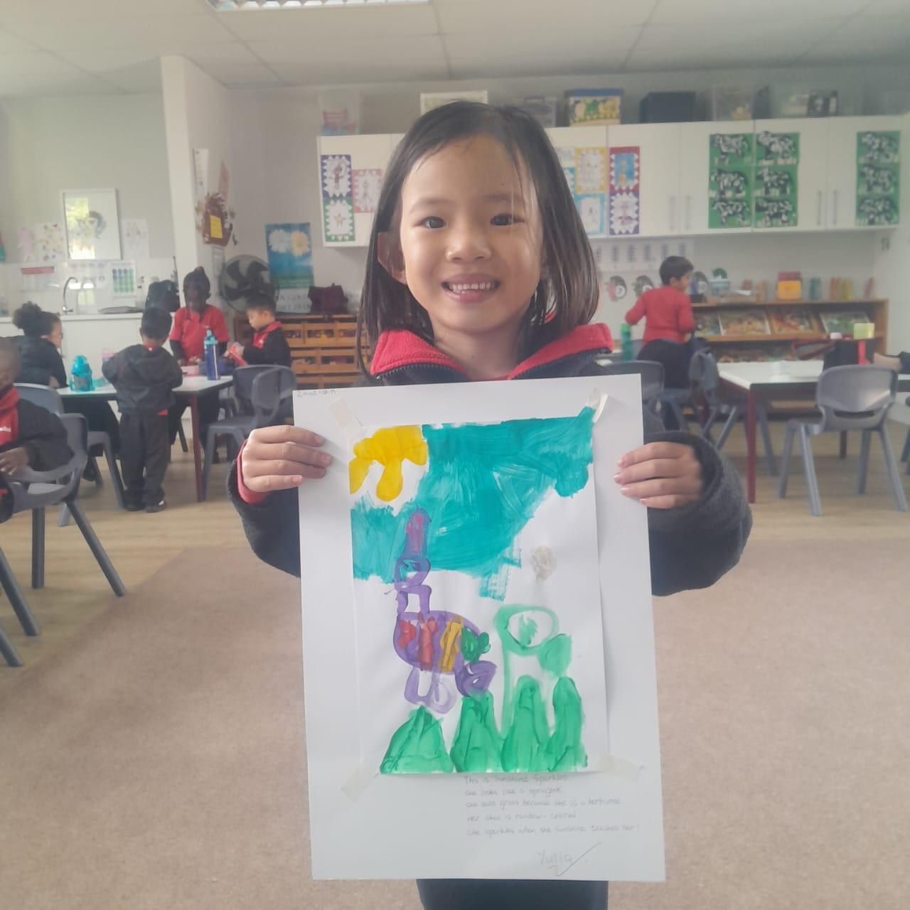 Girl holding up her colorful artwork in a classroom. The painting features green, yellow, and blue hues.