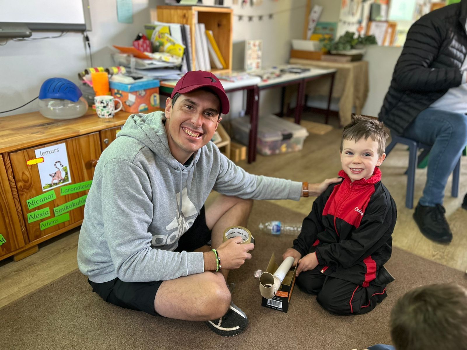 Man and child smiling on the floor. Man has arm around child. Cardboard toy cars are between them in a classroom setting.