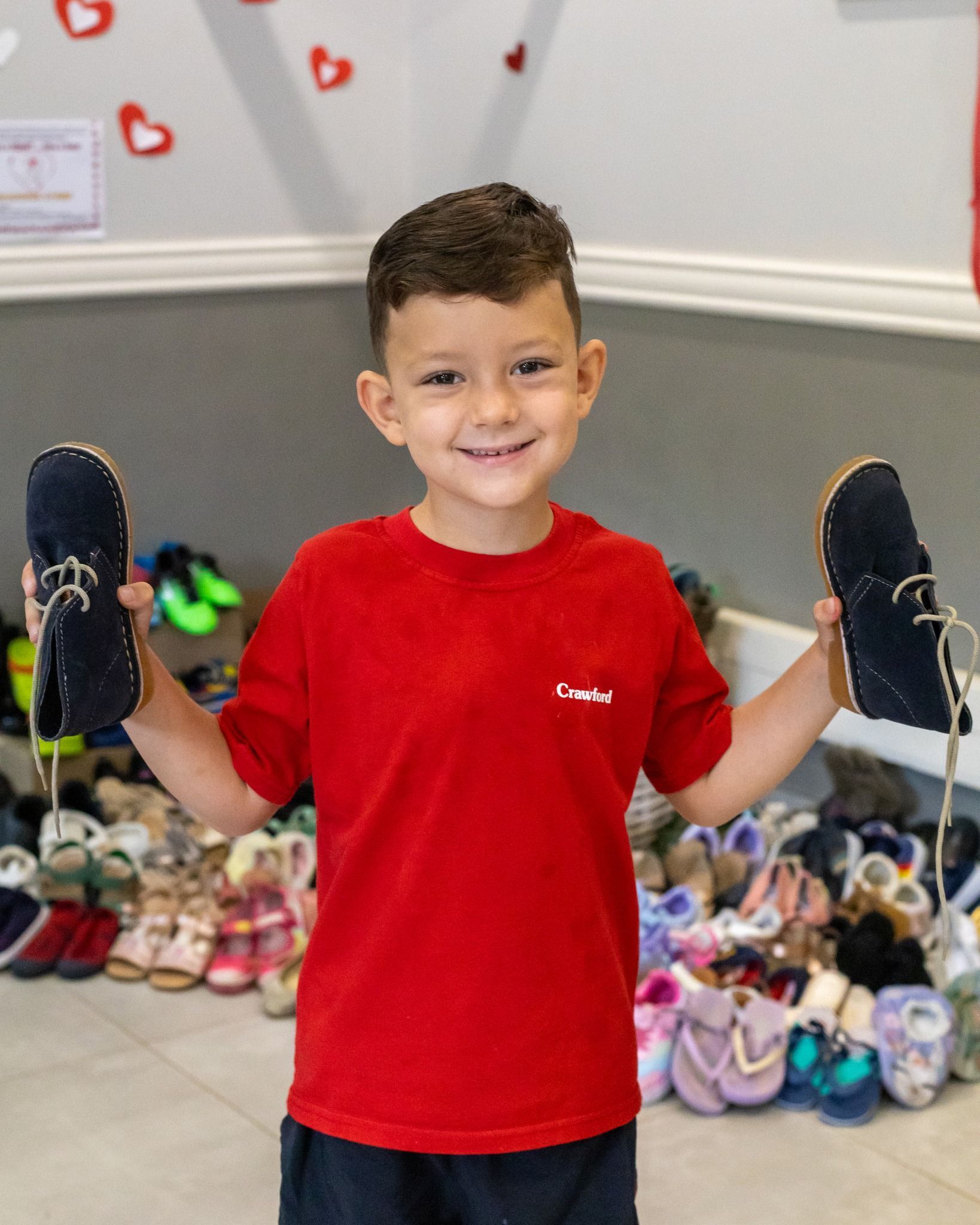 A young boy in a red shirt is holding two shoes in front of a pile of shoes.