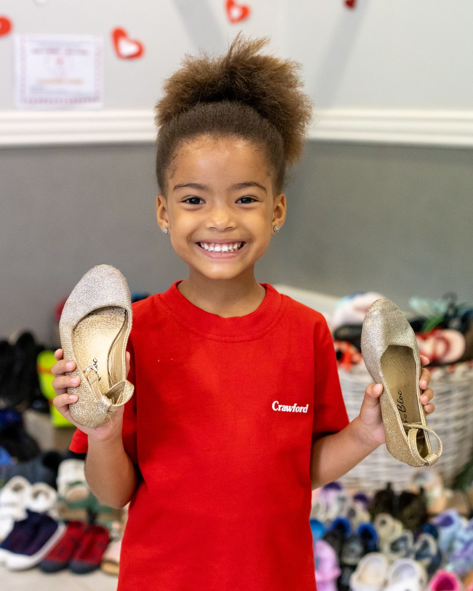 A little girl in a red shirt is holding a pair of shoes.