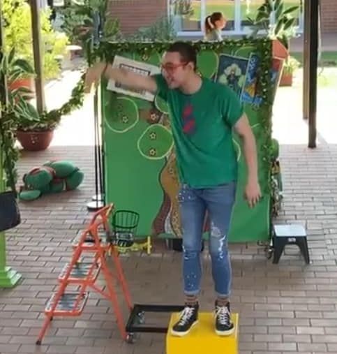 A man in a green shirt is standing on a yellow box in front of a green wall.