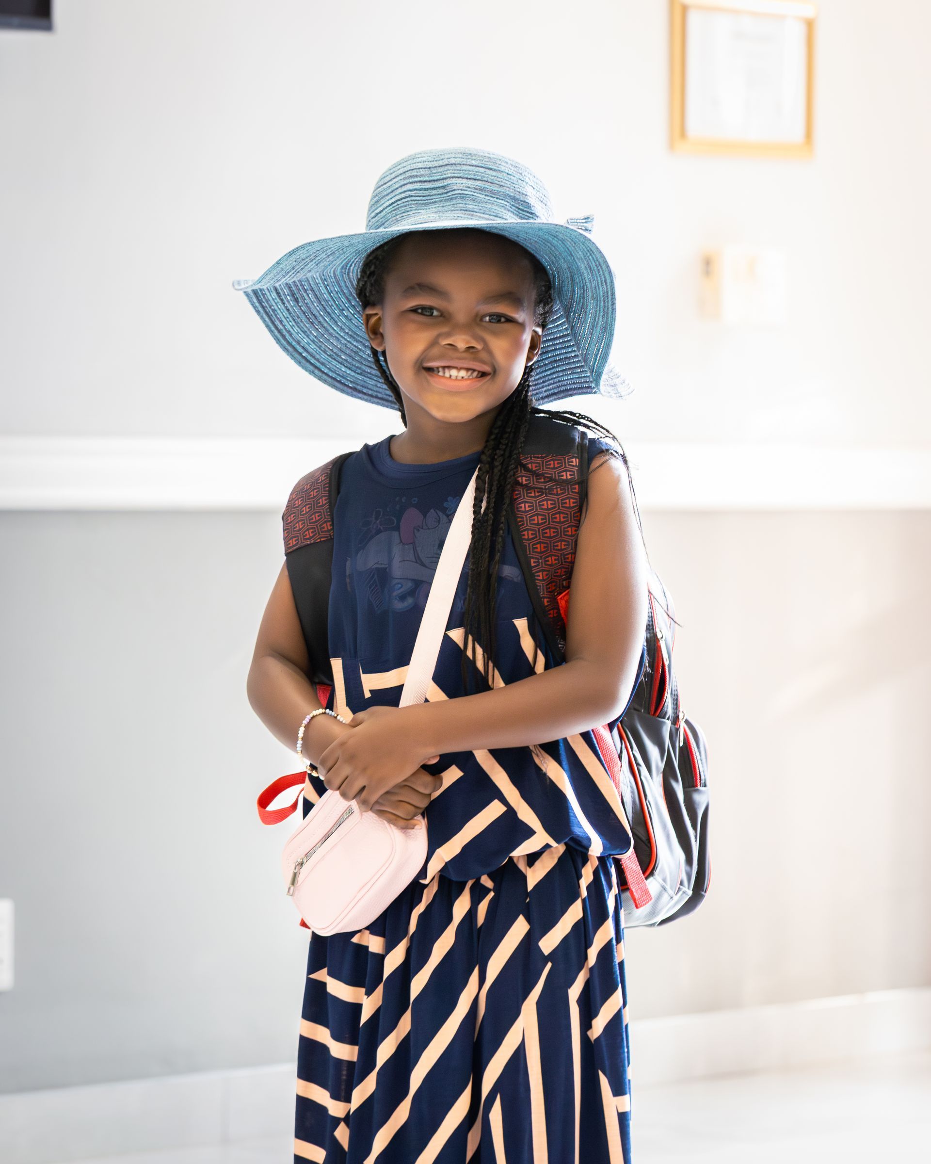 Girl wearing a blue hat, carrying a backpack and purse, smiling. Indoors.