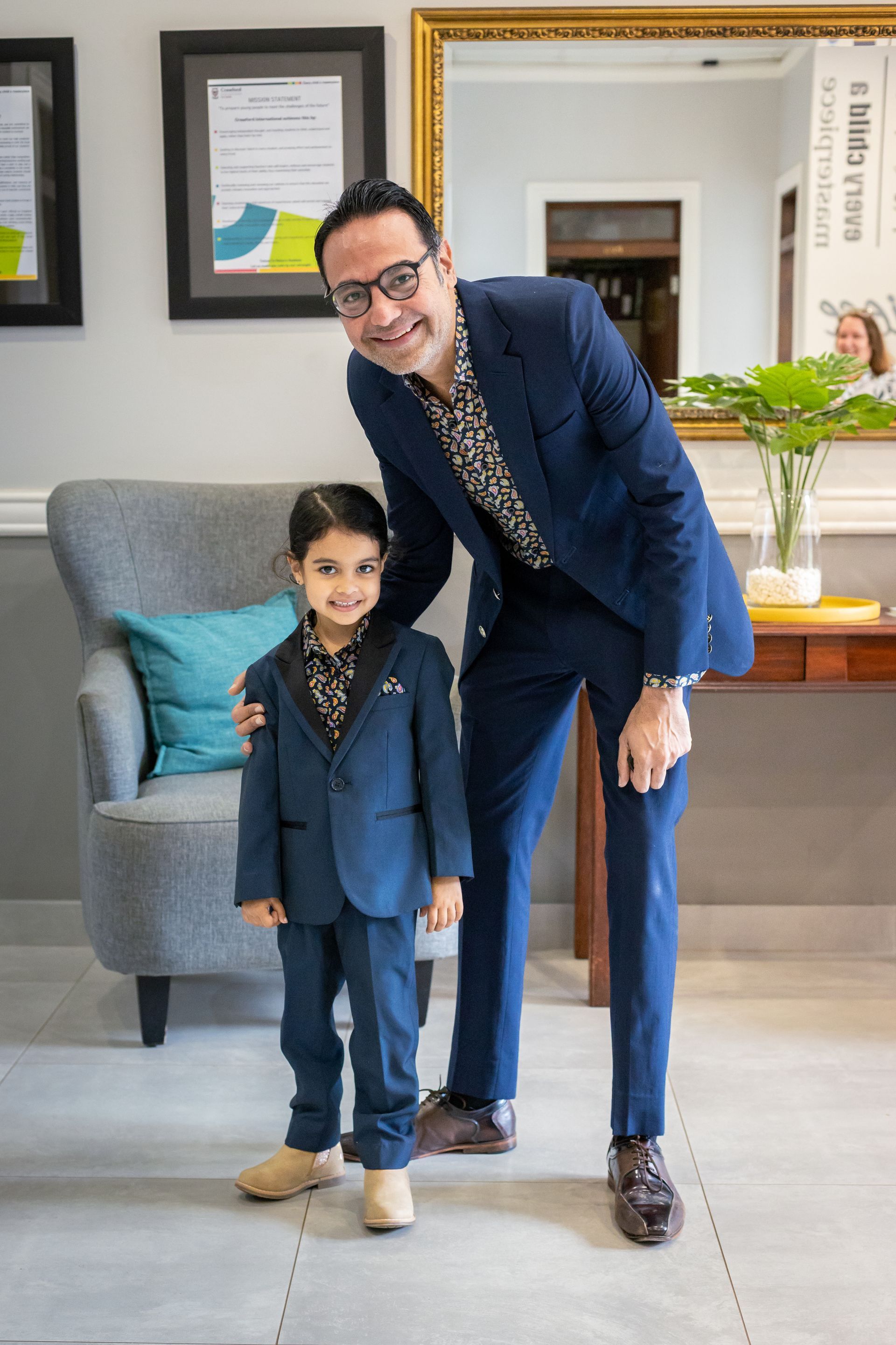A man and a child in suits smile; they stand near a chair and table in a room.