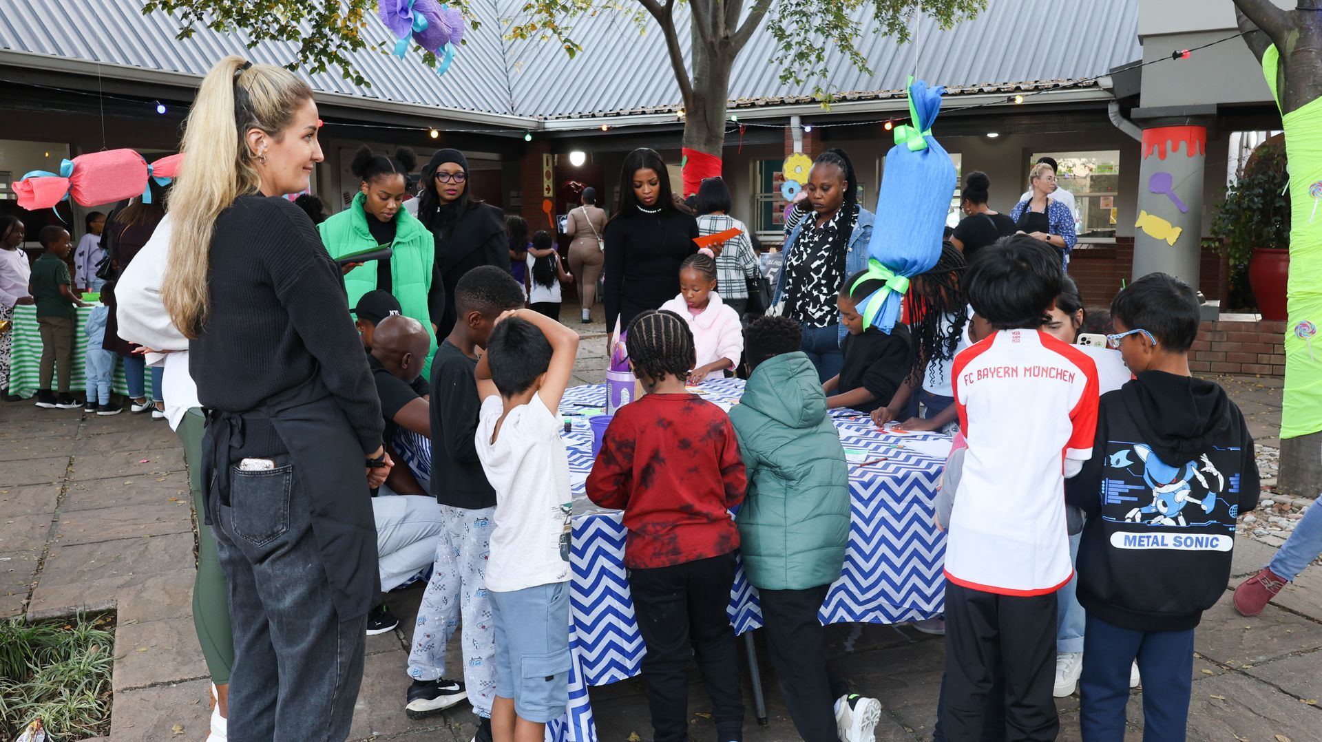 People gather around tables with children, possibly an event in an outdoor space.