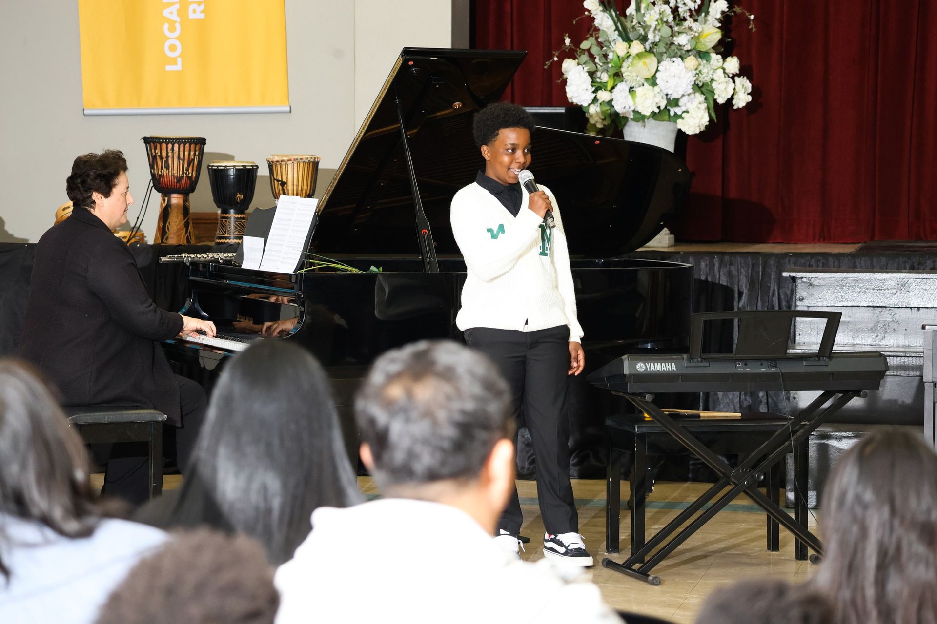 A person singing into a microphone on stage, with a piano player and audience present.