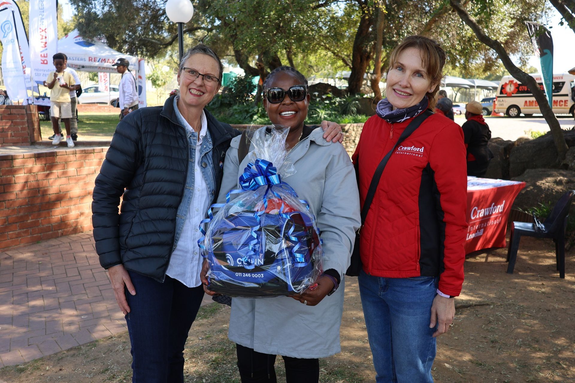 Three people smiling, one holding a gift basket, outdoors near tables and trees.