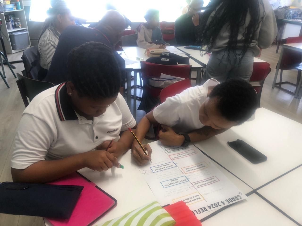 Two children are sitting at a table writing on a piece of paper.