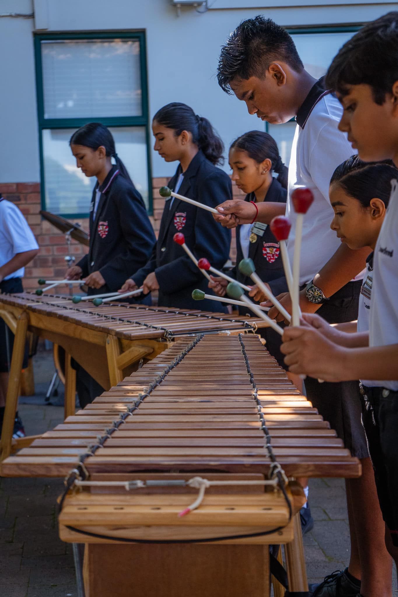 Children playing marimba outdoors, holding mallets. Several are wearing school uniforms.