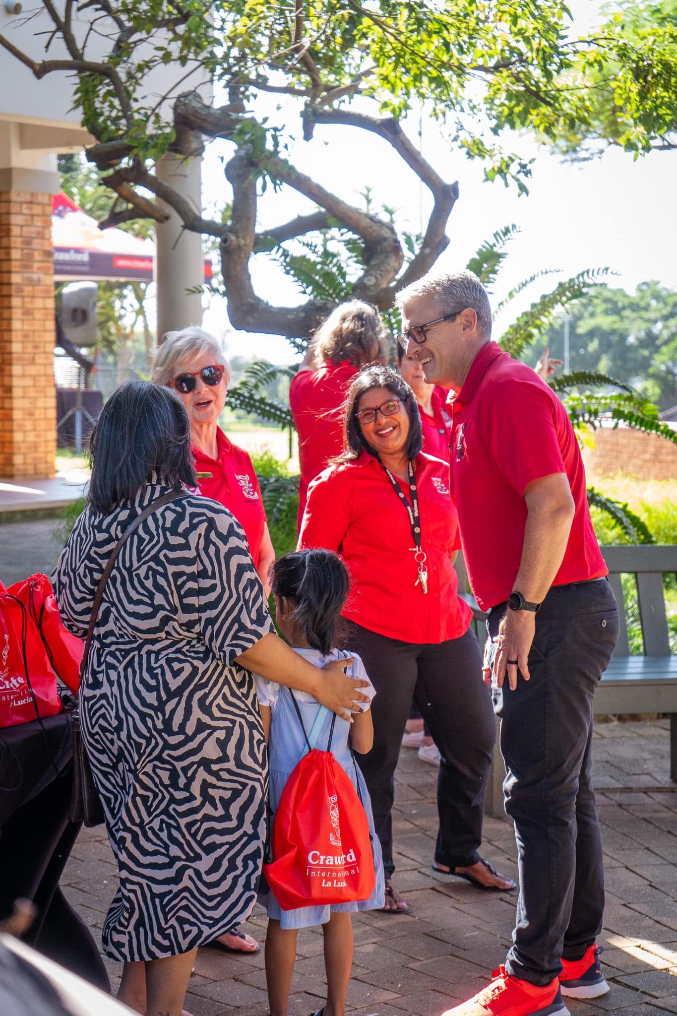 People in red shirts greeting others outside a building; a child in a blue dress hugs a woman.
