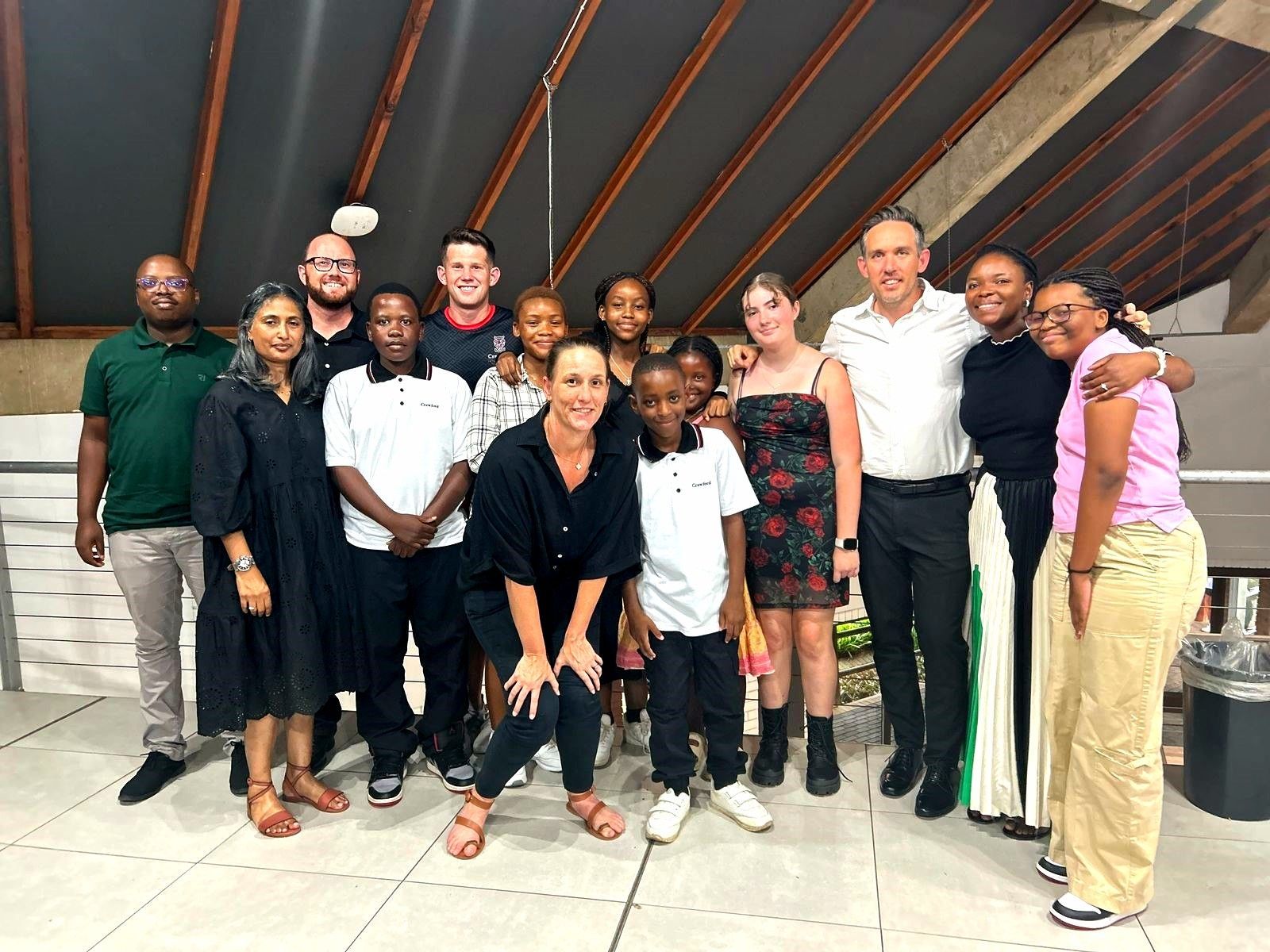 Group of people posing for photo indoors. Men, women, and children smiling, arms around each other.