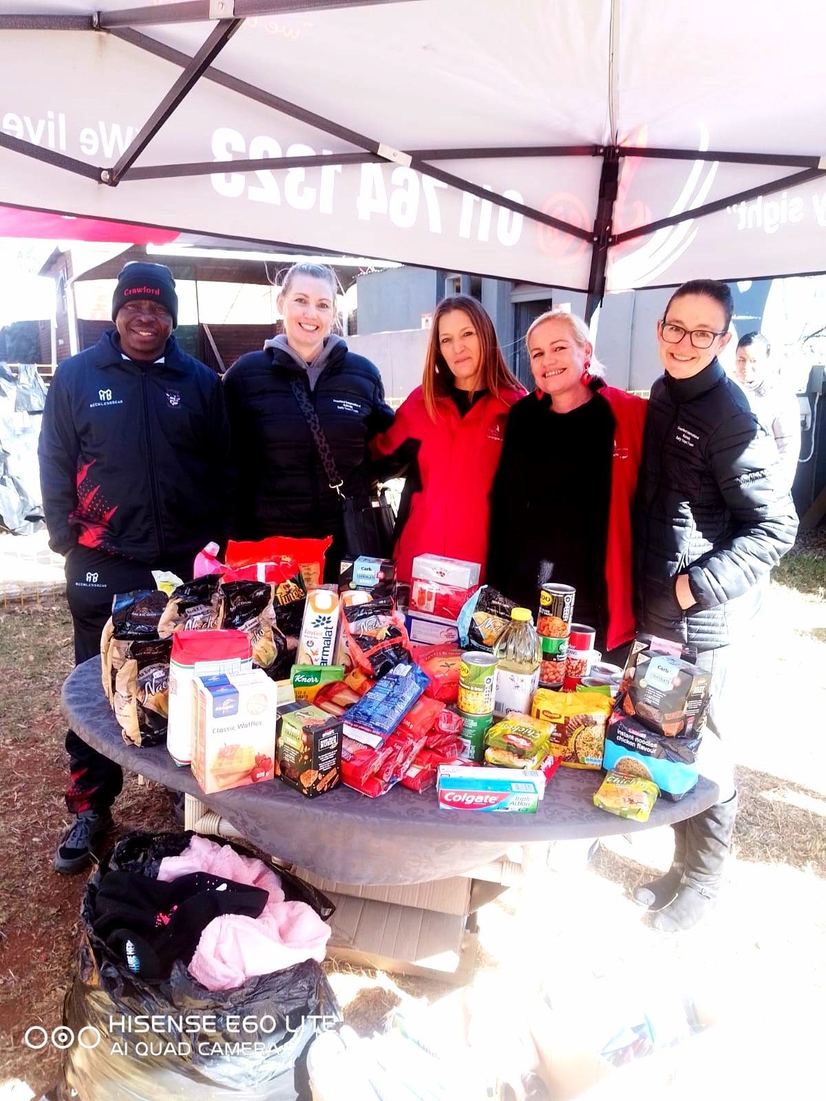 Five people pose behind a table laden with food items. They appear to be at an outdoor event under a tent.