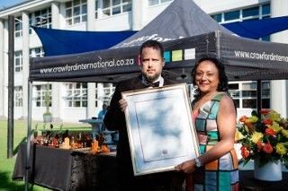 A man and woman are holding a framed picture in front of a tent that says crowford international.co.za