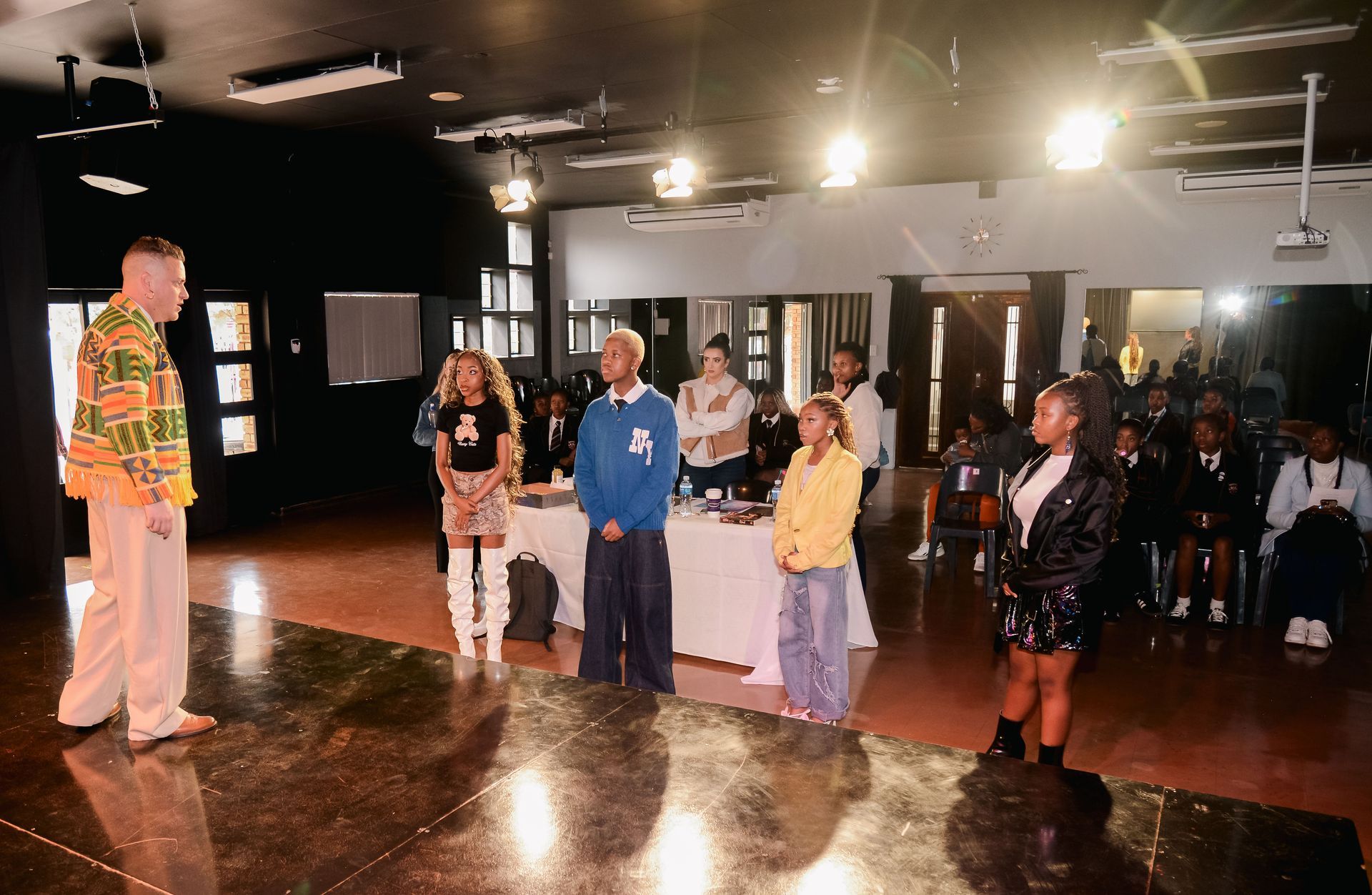 People standing on stage at event, tables behind them, audience in background, lights.