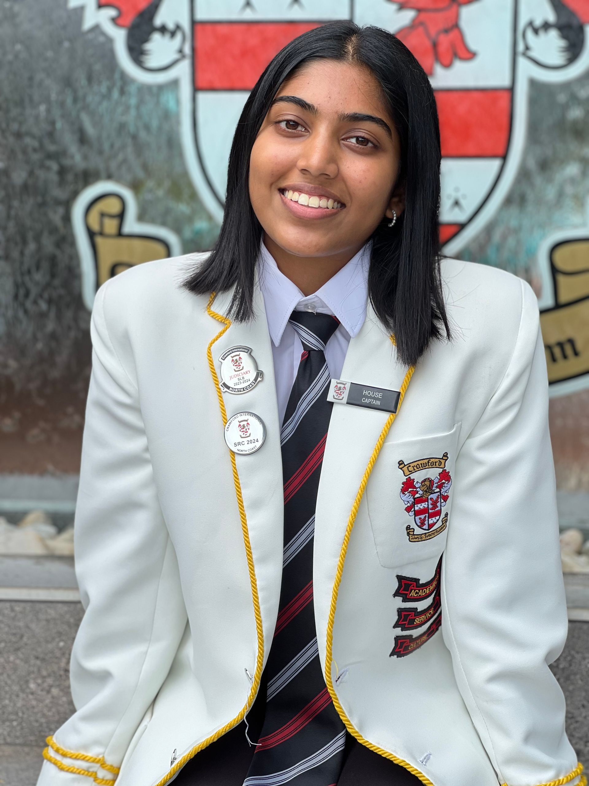 A young woman wearing a white jacket and tie is smiling in front of a coat of arms.