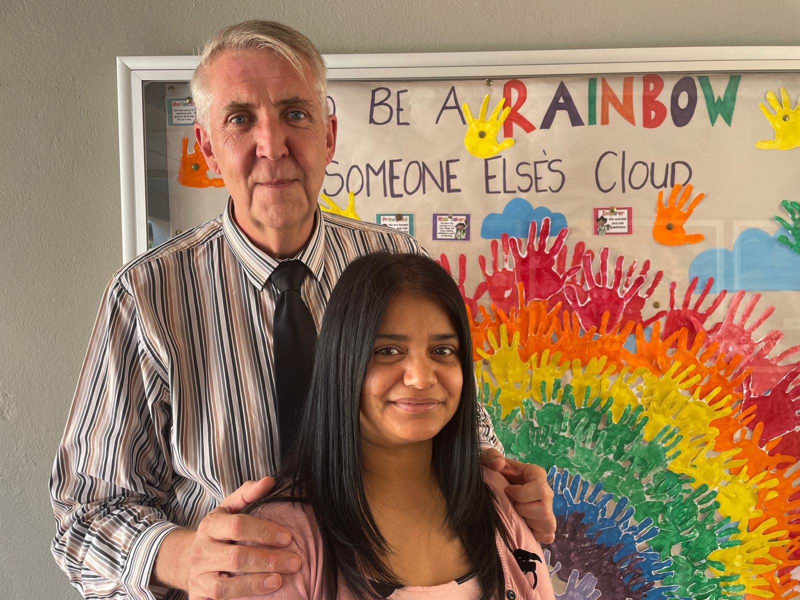 A man and a woman are posing in front of a sign that says be a rainbow someone else 's cloud.