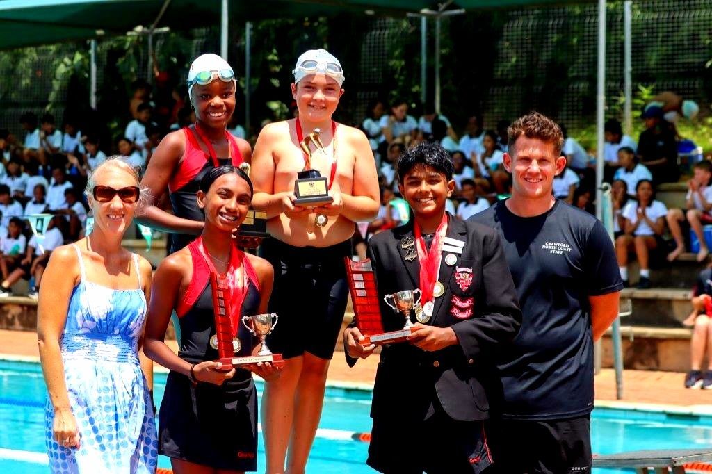 Swimmers stand on a pool deck, holding trophies and medals. A crowd watches in the background.