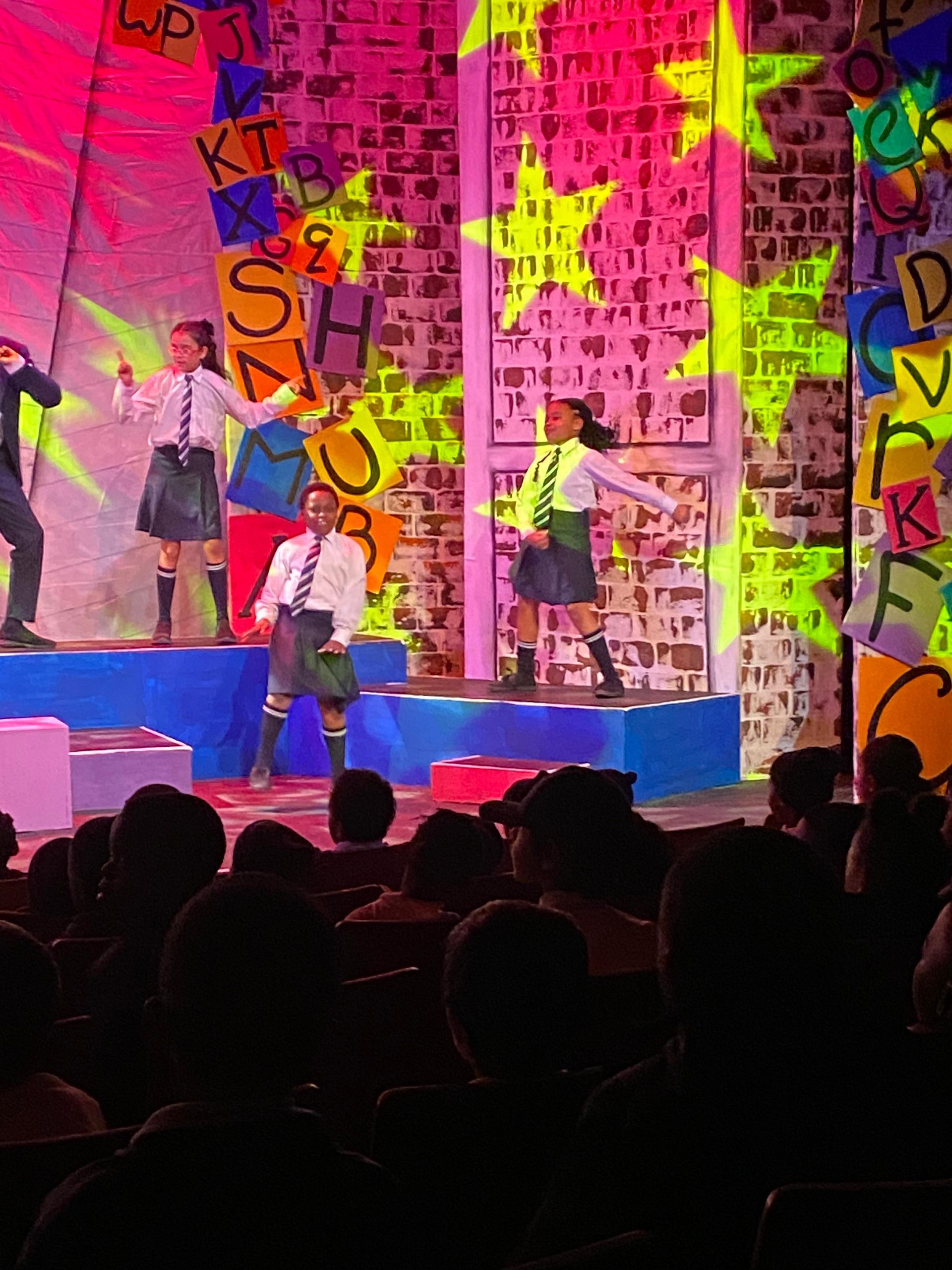 Children performing a play onstage with colorful set and audience.