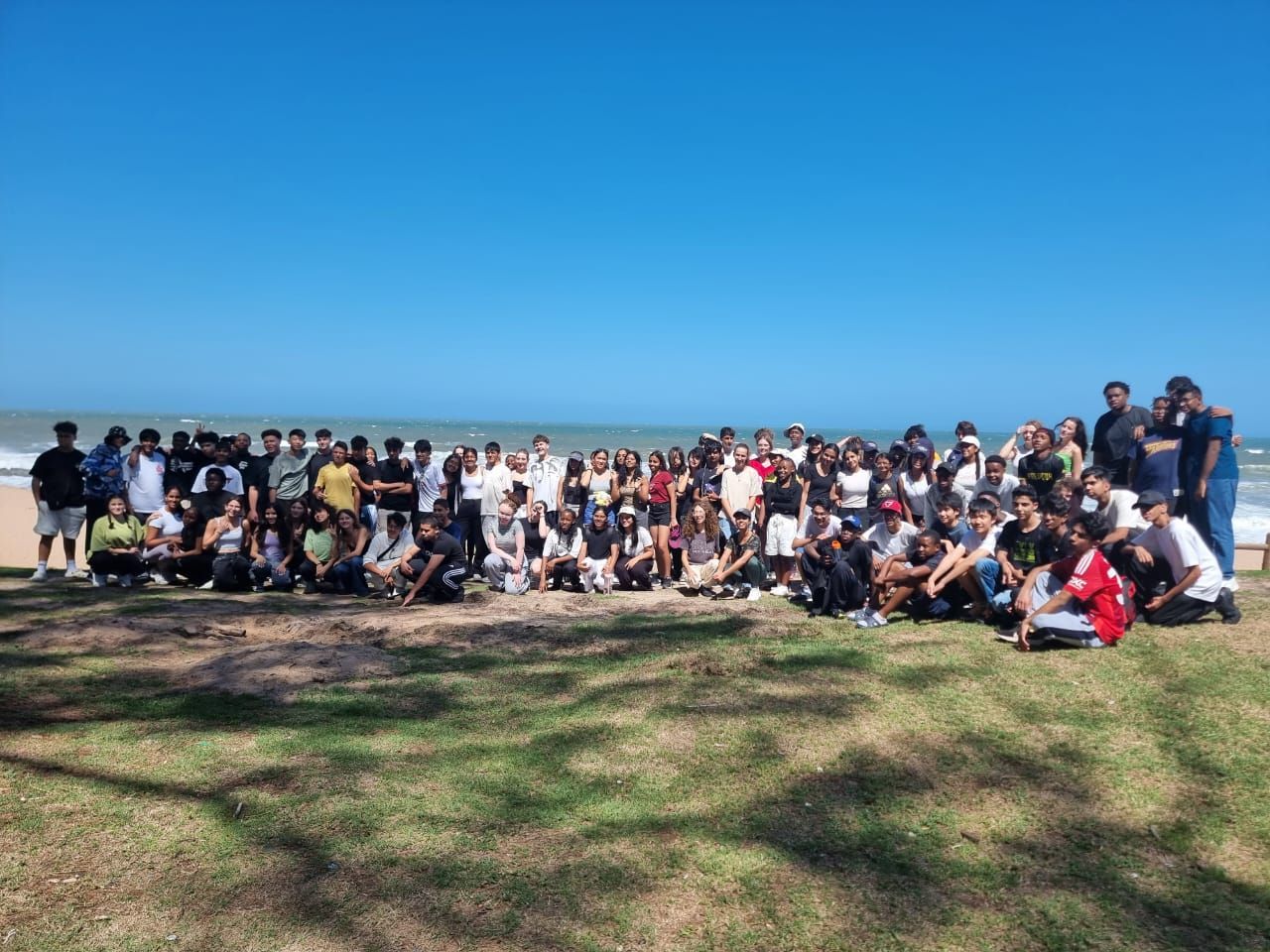 Group of people posing on a grassy area near the beach with clear blue sky.